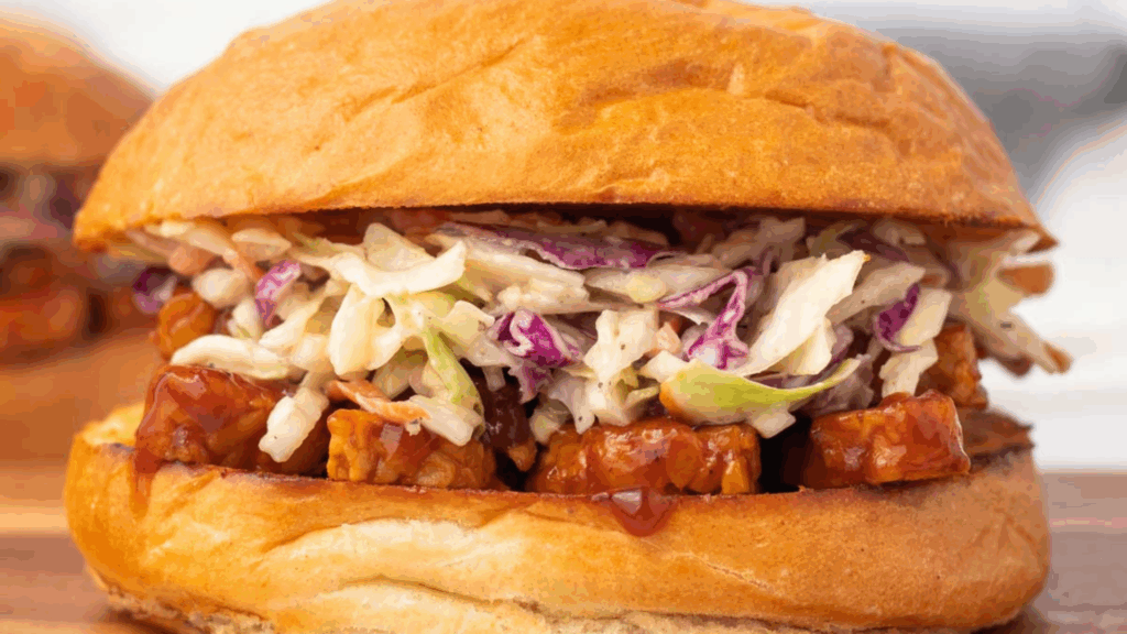 Sanduíche Plant-Based de Tempeh ao Barbecue com Salada de Abacaxi 1 Uma explosão de sabores defumados e tropicais em um sanduíche nutritivo e funcional Este sanduíche combina a textura firme do tempeh com cogumelos e grãos integrais, tudo envolto em um molho barbecue caseiro. A salada crocante de repolho roxo e abacaxi traz frescor e equilíbrio, tornando esta refeição perfeita para um almoço leve ou um jantar descontraído.
